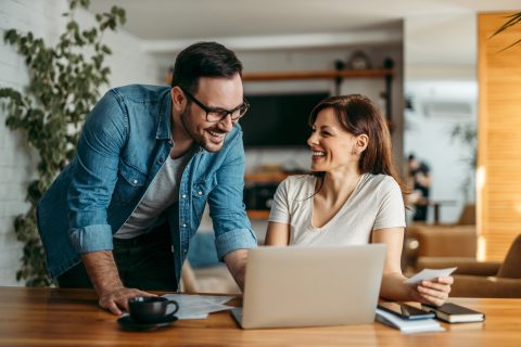 Great results! Portrait of a beautiful smiling woman sitting at desk with laptop and her coworker standing and looking at it, in the office.